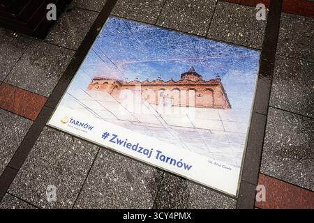 Tarnow, Polonia - 10 ottobre 2025: Una piastrella marciapiede che promuove Tarnów con un'immagine storica dell'edificio e un hashtag di viaggio. Foto Stock