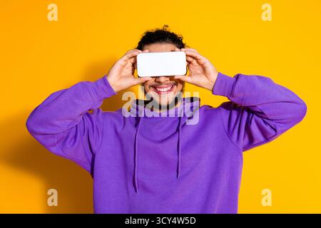 Uomo sorridente che posa su sfondo giallo con il telefono in mano con cappuccio viola per promuovere lo stile di vita moderno e il divertimento Foto Stock