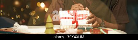 Uomo nero di mezza età che avvolge la confezione regalo di Natale con nastro rosso seduto a tavola, mani legate arco, decorazioni natalizie e luci dell'albero di Natale visibili sullo sfondo Foto Stock
