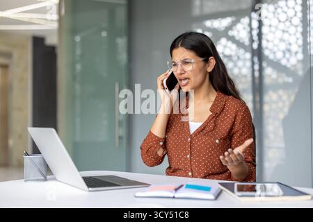 Giovane donna d'affari indiana che ha una conversazione telefonica intensa e arrabbiata mentre è seduta alla sua scrivania, sperimentando conflitti e stress in un ambiente di ufficio moderno Foto Stock