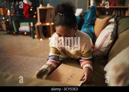 Bambina di colore sdraiata sul divano disegnando su carta immagini a tema natalizio con pennarelli colorati, luci festive e decorazioni natalizie visibili sullo sfondo, incentrate sull'attività creativa Foto Stock