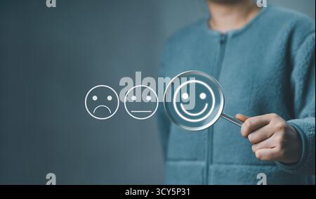 Analisi dei clienti feedback sulla soddisfazione del cliente. Mano con una lente d'ingrandimento con emoticon positivi tra neutri e negativi Foto Stock