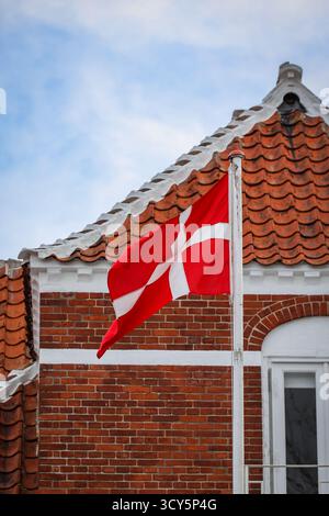 Bandiera danese che sventola di fronte a un tradizionale edificio in mattoni rossi con dettagli bianchi e tetto piastrellato a Skagen, Danimarca. Foto Stock