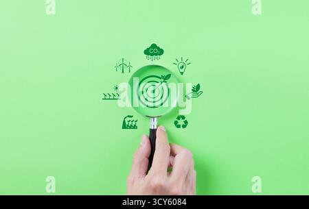 Energia verde e sostenibilità. Mano con una lente d'ingrandimento che mette a fuoco un bersaglio con una foglia, circondata da energia verde Foto Stock