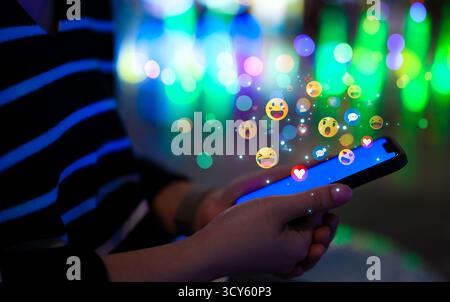 Concetto di tecnologia dei social media. Donna che utilizza uno smartphone con varie emoji colorate che fluttuano sopra lo schermo, simboleggiando l'interazione con i social media Foto Stock