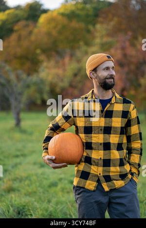 Ritratto di un giovane con la barba in un cappello arancione in un paesaggio autunnale. tenendo in mano una grande zucca. Foto Stock