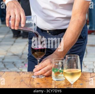 Primo piano di un uomo che versa vino rosso in un bicchiere su un tavolo di legno accanto ad altri bicchieri di vino bianco durante un evento di degustazione all'aperto. Foto Stock