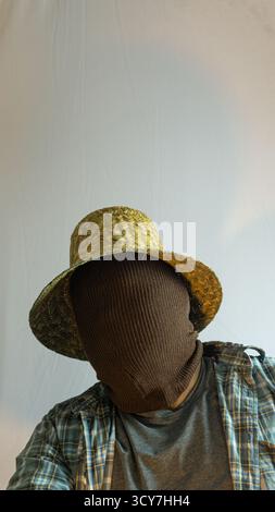 Primo piano di un agricoltore mascherato che indossa un cappello di paglia, viso completamente coperto di tessuto marrone, sfondo bianco che enfatizza il mistero e l'identità agricola Foto Stock