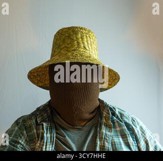Primo piano di un agricoltore mascherato che indossa un cappello di paglia, viso completamente coperto di tessuto marrone, sfondo bianco che enfatizza il mistero e l'identità agricola Foto Stock