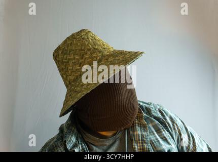 Primo piano di un agricoltore mascherato che indossa un cappello di paglia, viso completamente coperto di tessuto marrone, sfondo bianco che enfatizza il mistero e l'identità agricola Foto Stock
