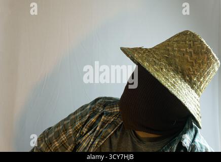 Primo piano di un agricoltore mascherato che indossa un cappello di paglia, viso completamente coperto di tessuto marrone, sfondo bianco che enfatizza il mistero e l'identità agricola Foto Stock