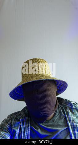 Primo piano di un agricoltore mascherato con cappello di paglia sotto la luce blu dell'ambiente, volto invisibile, carattere rurale con illuminazione stilizzata su sfondo bianco semplice Foto Stock