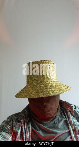 Contadino mascherato in cappello di paglia con volto nascosto, luce rossa aggiunge un tono drammatico al ritratto rurale, sfondo bianco minimalista con un'atmosfera intensa Foto Stock