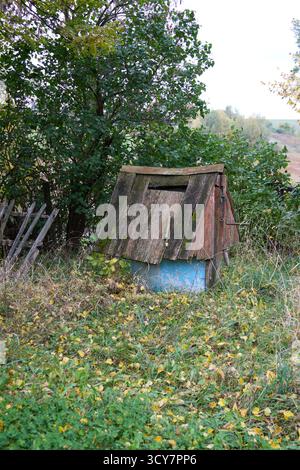 Antico pozzo con tetto in legno nel giardino autunnale, coperto di muschio, circondato da alberi e foglie gialle, un paesaggio rustico, base blu e pannelli in legno. Foto Stock