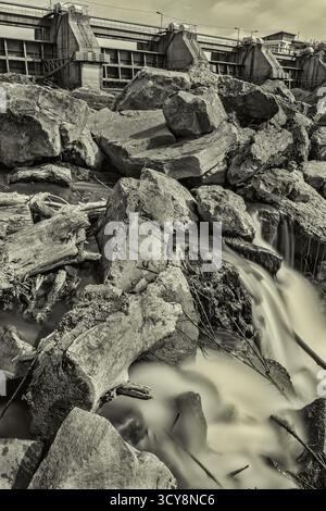 Lunga esposizione di acqua che scorre sopra le rocce vicino alla diga rumena. Le tonalità monocromatiche evidenziano il contrasto tra natura e ingegneria. Foto Stock
