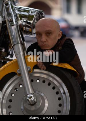 Primo piano di un uomo premuroso con nanismo seduto sulla ruota anteriore di una grande motocicletta da crociera gialla e guardando la telecamera. Chin riposa sulle mani, si concentra Foto Stock