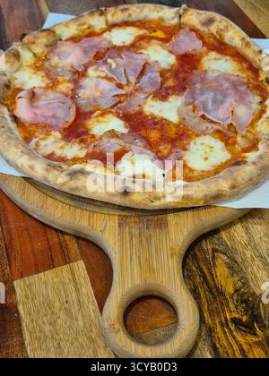 La foto mostra un primo piano di una pizza appena sfornata con prosciutto e formaggio, servita su una tavola rotonda di legno con manico Foto Stock