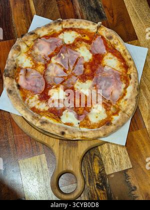 La foto mostra un primo piano di una pizza appena sfornata con prosciutto e formaggio, servita su una tavola rotonda di legno con manico Foto Stock