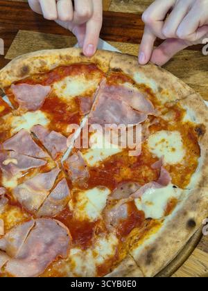 La foto mostra un primo piano di una pizza appena sfornata con prosciutto e formaggio, servita su una tavola rotonda di legno con manico Foto Stock