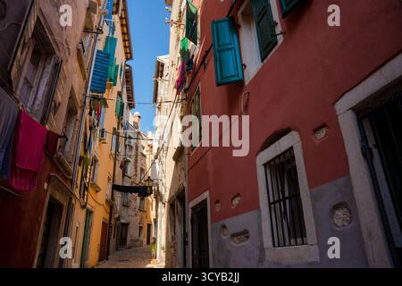 Rovigno, Croazia - 15 agosto 2025: Vicolo stretto a Rovigno - vecchi edifici colorati con persiane aperte e lavanderia appesa su un ciottolo illuminato dal sole Foto Stock