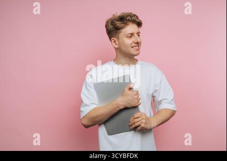 Un giovane si trova con sicurezza contro un muro rosa, tenendo un portatile vicino al petto. Sorride e sembra rilassato, vestito casualmente in un bianco Foto Stock