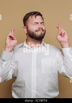 Un uomo con la barba sta in piedi su uno sfondo neutrale, incrociando le dita con un'espressione di speranza. Indossa una camicia bianca e ha la sua Foto Stock