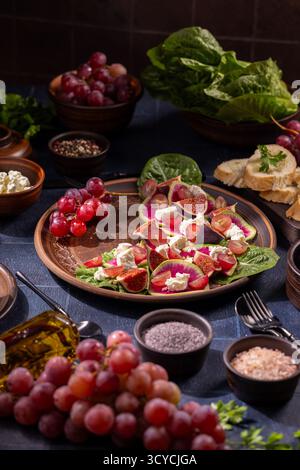 Vivace insalata mediterranea con fichi freschi, uva, feta sbriciolata e ravanello anguria sulle verdure, piatto rustico in ceramica con olio d'oliva e condimenti Foto Stock