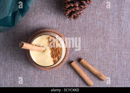 Un bicchiere di crema, classico eggnog guarnito con un bastoncino di cannella e una spolverata pesante di noce moscata in polvere, fotografato da una vista dall'alto su un sacco di sughero Foto Stock