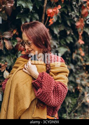 Donna con sciarpa gialla senape e maglione testurizzato rosso con capelli intrecciati in piedi vicino alla parete edera morbido umore autunnale accogliente stile di vita lento tranquillo Foto Stock