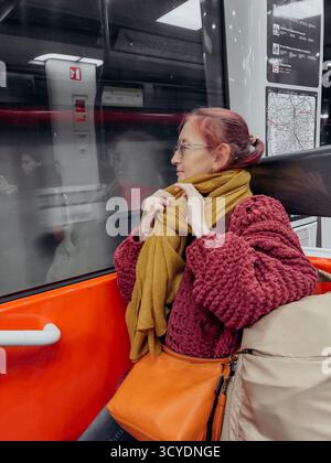 Donna con borsa arancione e sciarpa di senape seduta in tram che guarda fuori dalla finestra i riflessi sfocati indossando un maglione rosso testurizzato atmosfera introspettiva Foto Stock