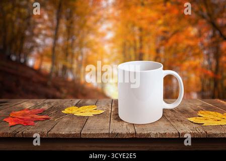 Tazza da caffè bianca su un tavolo rustico con foglie autunnali colorate e sfondo boschivo sfocato, ideale per i modelli di design Foto Stock