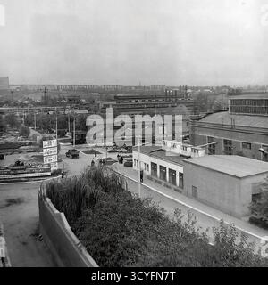 Questa foto archivistica in bianco e nero degli anni '1970 offre una vista panoramica ad alto angolo dell'impianto isolatore armatura di Slavyansk (AIZ) a Sloviansk, URSS. Lo scatto cattura il vasto paesaggio industriale, mostrando varie officine, strade interne e infrastrutture. Nella vista, i lavoratori camminano lungo i sentieri e sono visibili grandi cartelloni pubblicitari (Doska Pocheta). L'ampio complesso comprende magazzini all'aperto con prodotti finiti, binari ferroviari per la logistica e camini industriali alti. Sullo sfondo lontano, la fabbrica si fonde con lo skyline residenziale della città. Foto Stock