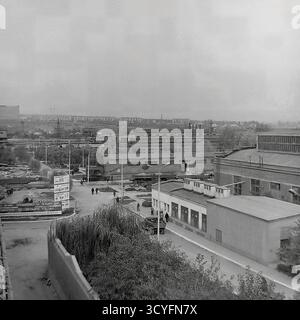 Questa foto archivistica in bianco e nero degli anni '1970 offre una vista panoramica ad alto angolo dell'impianto isolatore armatura di Slavyansk (AIZ) a Sloviansk, URSS. Lo scatto cattura il vasto paesaggio industriale, mostrando varie officine, strade interne e infrastrutture. Nella vista, i lavoratori camminano lungo i sentieri e sono visibili grandi cartelloni pubblicitari (Doska Pocheta). L'ampio complesso comprende magazzini all'aperto con prodotti finiti, binari ferroviari per la logistica e camini industriali alti. Sullo sfondo lontano, la fabbrica si fonde con lo skyline residenziale della città. Foto Stock