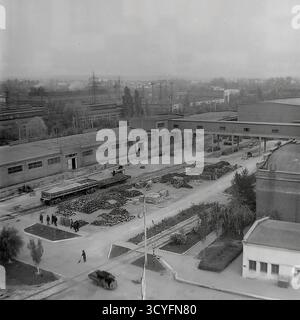 Questa foto archivistica in bianco e nero degli anni '1970 offre una vista panoramica ad alto angolo dell'impianto isolatore armatura di Slavyansk (AIZ) a Sloviansk, URSS. Lo scatto cattura il vasto paesaggio industriale, mostrando varie officine, strade interne e infrastrutture. Nella vista, i lavoratori camminano lungo i sentieri e sono visibili grandi cartelloni pubblicitari (Doska Pocheta). L'ampio complesso comprende magazzini all'aperto con prodotti finiti, binari ferroviari per la logistica e camini industriali alti. Sullo sfondo lontano, la fabbrica si fonde con lo skyline residenziale della città. Foto Stock