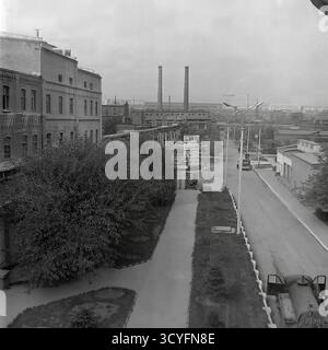 Questa foto archivistica in bianco e nero degli anni '1970 offre una vista panoramica ad alto angolo dell'impianto isolatore armatura di Slavyansk (AIZ) a Sloviansk, URSS. Lo scatto cattura il vasto paesaggio industriale, mostrando varie officine, strade interne e infrastrutture. Nella vista, i lavoratori camminano lungo i sentieri e sono visibili grandi cartelloni pubblicitari (Doska Pocheta). L'ampio complesso comprende magazzini all'aperto con prodotti finiti, binari ferroviari per la logistica e camini industriali alti. Sullo sfondo lontano, la fabbrica si fonde con lo skyline residenziale della città. Foto Stock