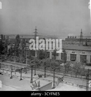 Questa foto archivistica in bianco e nero degli anni '1970 offre una vista panoramica ad alto angolo dell'impianto isolatore armatura di Slavyansk (AIZ) a Sloviansk, URSS. La foto si affaccia sul tetto di un edificio su una lunga officina, alberi e il vasto paesaggio industriale. Sullo sfondo sono visibili numerosi piloni di trasmissione elettrica (torri di potenza), camini industriali e grandi serbatoi di stoccaggio. Questa immagine documenta la portata dell'industria sovietica Foto Stock