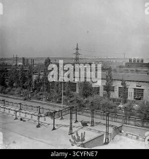 Questa foto archivistica in bianco e nero degli anni '1970 offre una vista panoramica ad alto angolo dell'impianto isolatore armatura di Slavyansk (AIZ) a Sloviansk, URSS. La foto si affaccia sul tetto di un edificio su una lunga officina, alberi e il vasto paesaggio industriale. Sullo sfondo sono visibili numerosi piloni di trasmissione elettrica (torri di potenza), camini industriali e grandi serbatoi di stoccaggio. Questa immagine documenta la portata dell'industria sovietica Foto Stock