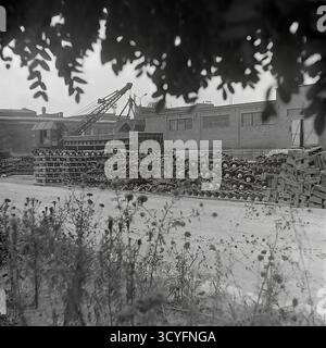 Questa foto d'archivio in bianco e nero degli anni '1970 di Sloviansk, URSS, mostra una vista dell'impianto isolatore armatura di Slavyansk (AIZ). La scena è incorniciata da foglie e piante sfocate in primo piano. Il cantiere industriale è visto come un grande magazzino all'aperto, pieno di enormi pile di isolanti finiti e pallet di legno vuoti. Sullo sfondo, una gru pesante montata su un carrello è parcheggiata accanto a un lungo edificio di officina in mattoni. Questa immagine quadrata documenta la scala di produzione nella fabbrica sovietica Foto Stock