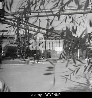 Questa foto d'archivio in bianco e nero degli anni '1970 di Sloviansk, URSS, mostra una vista dell'impianto isolatore armatura di Slavyansk (AIZ). Lo scatto viene inquadrato artisticamente attraverso rami di alberi sfocati. Cattura la complessa infrastruttura industriale, tra cui un grande serbatoio orizzontale (caldaia/cisterna) e una rete di condotte sopraelevate su capriate metalliche. Sullo sfondo sono visibili un'officina di mattoni e un poster di propaganda. Questa immagine documenta il paesaggio industriale sovietico. Foto Stock