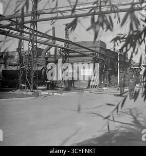 Questa foto d'archivio in bianco e nero degli anni '1970 di Sloviansk, URSS, mostra una vista dell'impianto isolatore armatura di Slavyansk (AIZ). Lo scatto viene inquadrato artisticamente attraverso rami di alberi sfocati. Cattura la complessa infrastruttura industriale, tra cui un grande serbatoio orizzontale (caldaia/cisterna) e una rete di condotte sopraelevate su capriate metalliche. Sullo sfondo sono visibili un'officina di mattoni e un poster di propaganda. Questa immagine documenta il paesaggio industriale sovietico. Foto Stock