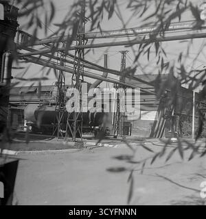 Questa foto d'archivio in bianco e nero degli anni '1970 di Sloviansk, URSS, mostra una vista dell'impianto isolatore armatura di Slavyansk (AIZ). Lo scatto viene inquadrato artisticamente attraverso rami di alberi sfocati. Cattura la complessa infrastruttura industriale, tra cui un grande serbatoio orizzontale (caldaia/cisterna) e una rete di condotte sopraelevate su capriate metalliche. Sullo sfondo sono visibili un'officina di mattoni e un poster di propaganda. Questa immagine documenta il paesaggio industriale sovietico. Foto Stock