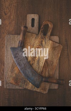 Un grande coltello ricurvo poggia su due taglieri in legno su una superficie rustica. Il coltello mostra usura, mentre le tavole mostrano un esclusivo tipo di granella Foto Stock