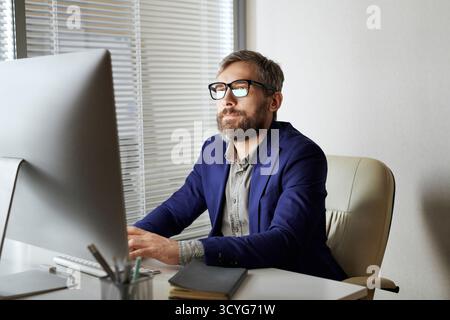 Uomo d'affari caucasico di mezza età con barba e bicchieri seduto su una comoda sedia e lavorando al computer in un ufficio moderno Foto Stock