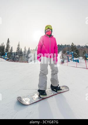 Uno snowboarder si erge su una pista innevata, vestito con una giacca rosa vibrante e occhiali di protezione Foto Stock