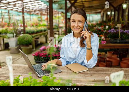 Un giovane professionista che lavora diligentemente in un negozio di fiori utilizzando sia un computer portatile che un telefono Foto Stock