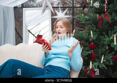 Sorpresa donna caucasica bionda che vince un premio online su smartphone vicino all'albero di Natale Foto Stock