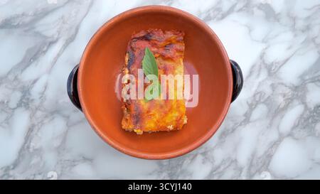 Lasagne fatte in casa con salsa di pomodoro e foglie di basilico servite in una rustica ciotola di terracotta su un tavolo di marmo. Foto Stock