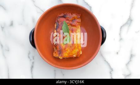 Lasagne fatte in casa con salsa di pomodoro e foglie di basilico servite in una rustica ciotola di terracotta su un tavolo di marmo. Foto Stock