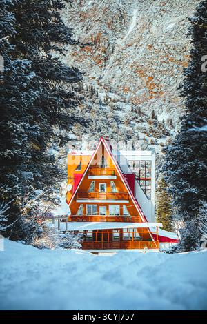 Vista ravvicinata di un vivace rifugio di montagna in stile A-frame rosso e arancione annidato in una foresta di conifere ricoperta di neve Foto Stock