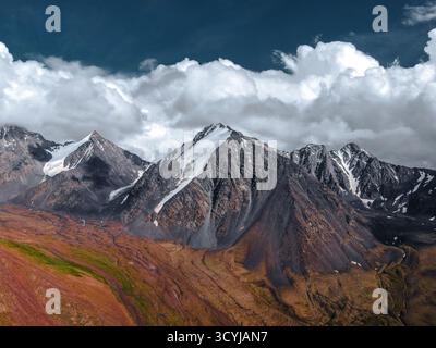 Vista aerea panoramica delle aspre cime di montagna e delle pendici con contrasti sorprendenti di rosso-marrone, roccia nera e neve bianca sotto un cielo nuvoloso Foto Stock
